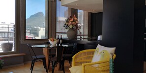 A modern dining area with a glass table, black chairs, and a yellow wicker chair. A vase with flowers is on the windowsill, and a bowl of fruit and wine is on the table. - Cartwrights Apartments (Cape Town)