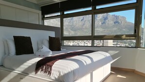 Ein modernes Schlafzimmer mit einem großen Bett, weißer Bettwäsche und Blick auf eine Stadtlandschaft und Berge durch große Fenster.