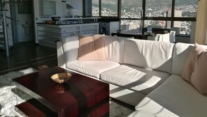 Ein modernes Wohnzimmer mit einem weißen Sofa, einem Holztisch und Blick auf eine Stadtlandschaft durch große Fenster.