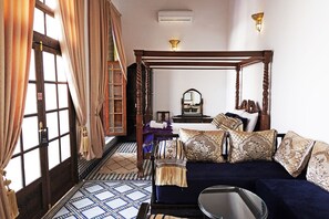 A traditional four-poster bed with a mirror above it, a sofa with patterned cushions, and a small round table.