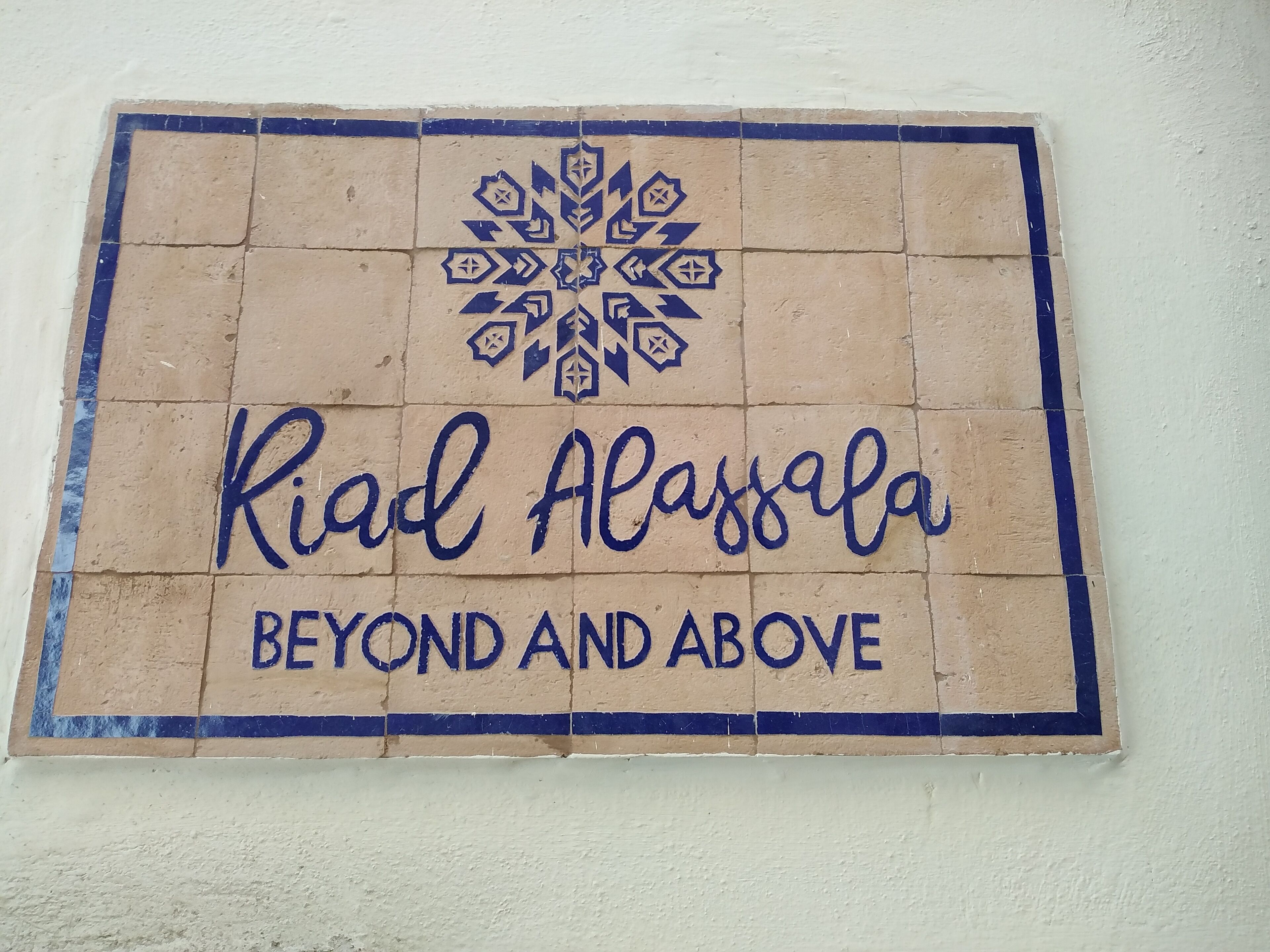 Een bord met de tekst 'Riad Alassafa' en 'BEYOND AND ABOVE' op een betegelde muur.