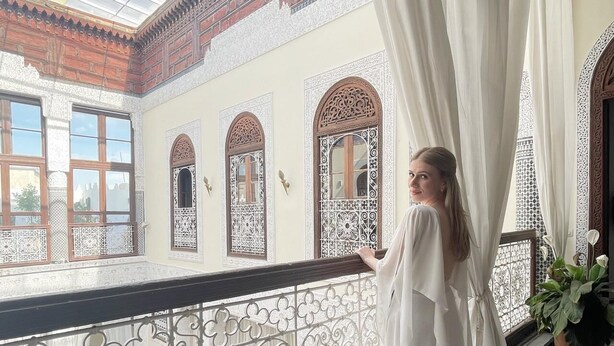 Una mujer con vestido blanco de pie en un balcón con barandillas ornamentales y baldosas estampadas.
