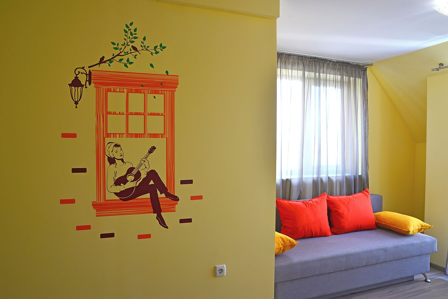 Una habitación con pared amarilla, ventana con cortinas traslúcidas, sofá gris con cojines rojo y amarillo, y un adhesivo mural de una persona tocando la guitarra.
