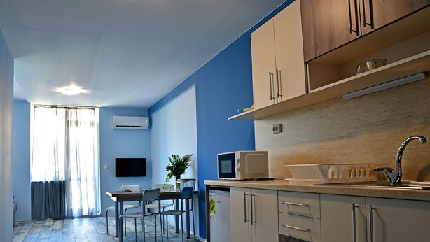 A modern kitchen with a microwave, sink, and cabinets. There is a dining area with chairs and a table. - Visito Apart House (Sofia)