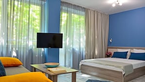 A modern hotel room with a large bed, a television, a small table with a bowl, and bright orange and blue pillows. - Visito Apart House (Sofia)
