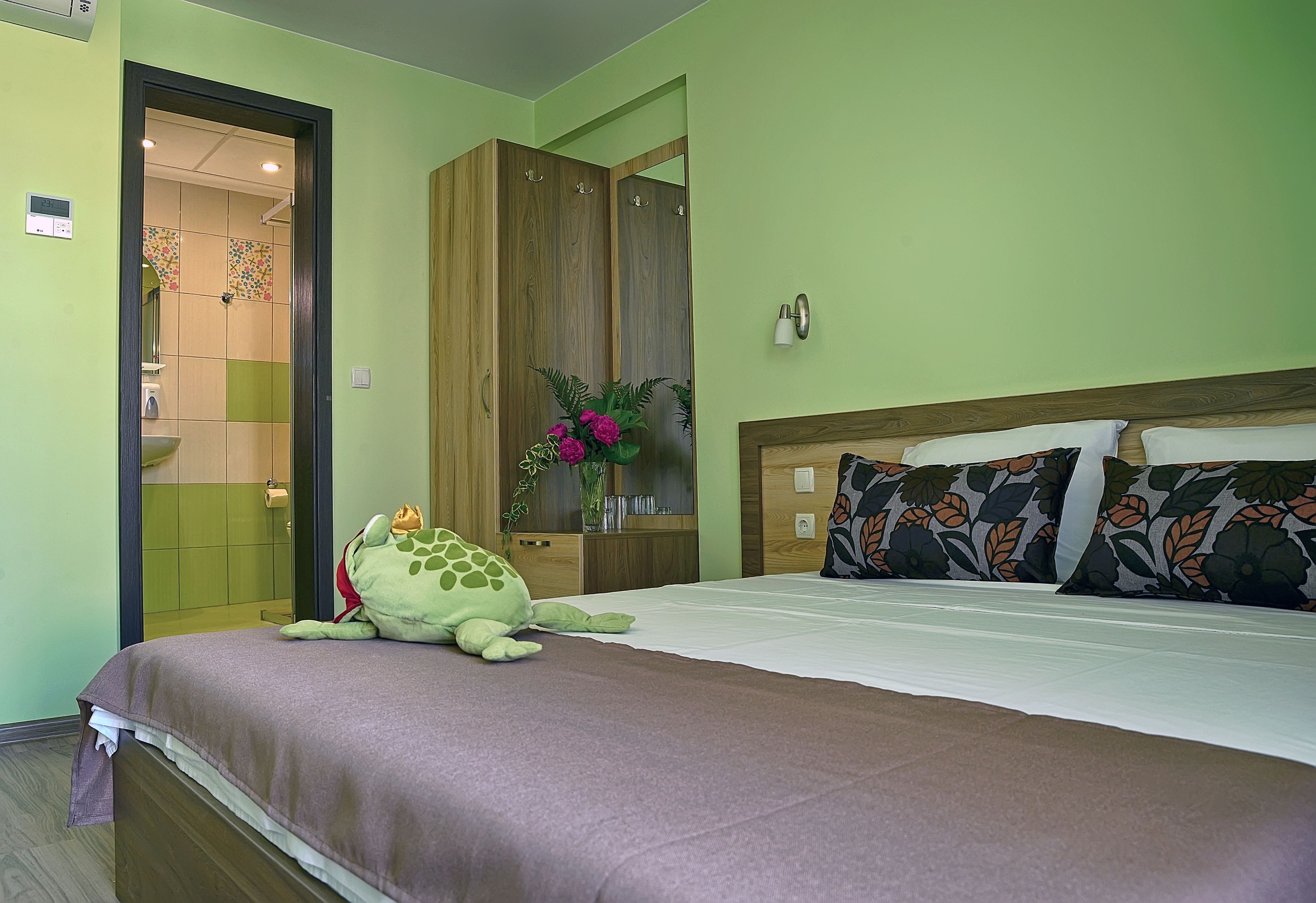 Habitación de hotel con cama, armario, baño visible a través de una puerta abierta y un peluche decorativo de rana sobre la cama.