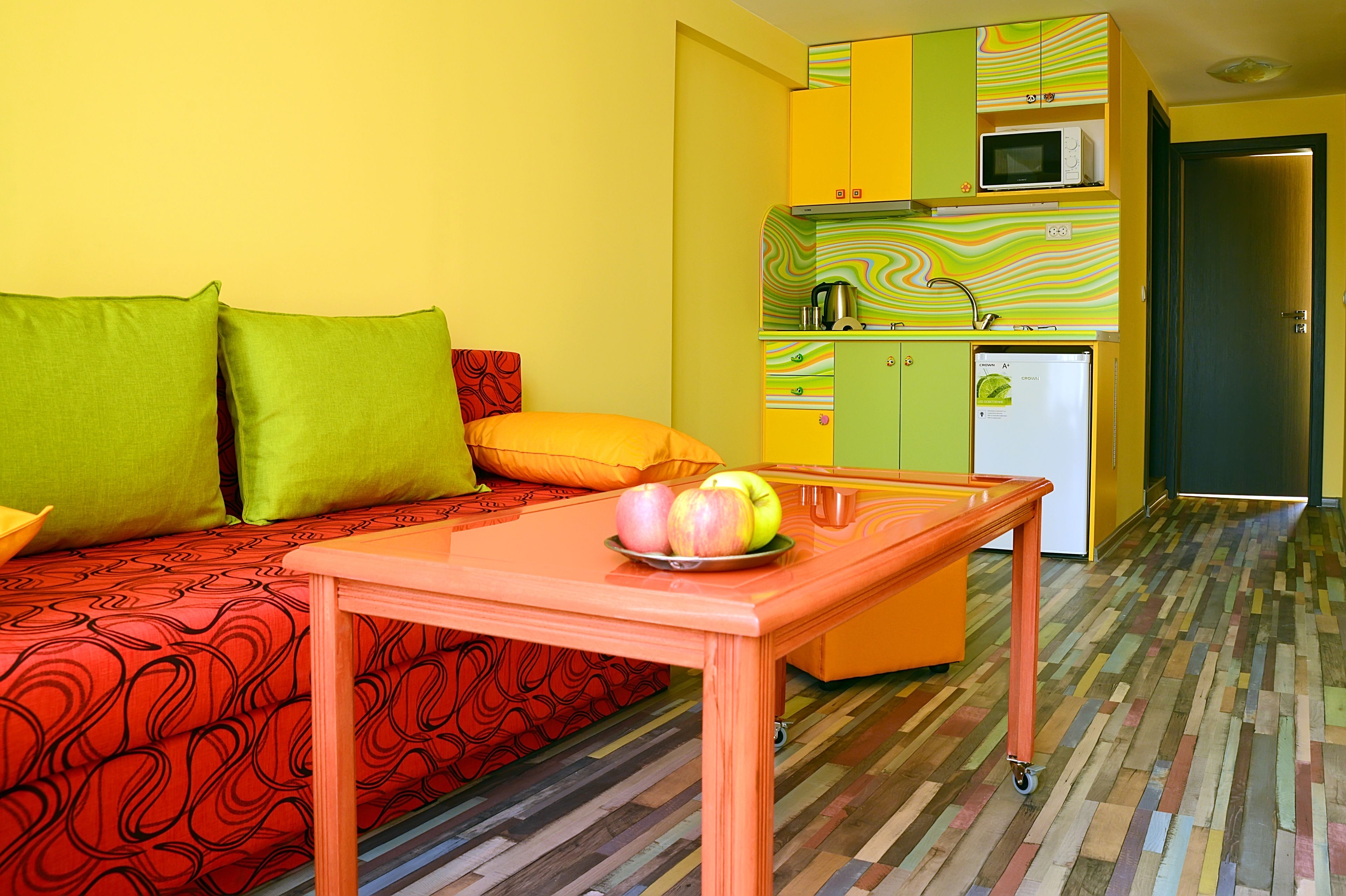A compact living space with a sofa, a small table with fruit, a kitchenette, and a door leading to another room.