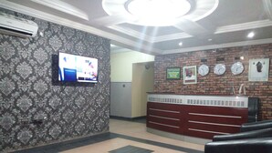 A reception area with a patterned wallpaper, a flat-screen TV, a brick wall with clocks, and a counter with drawers. - Akma Signature Hotel & Suites (Ibadan)