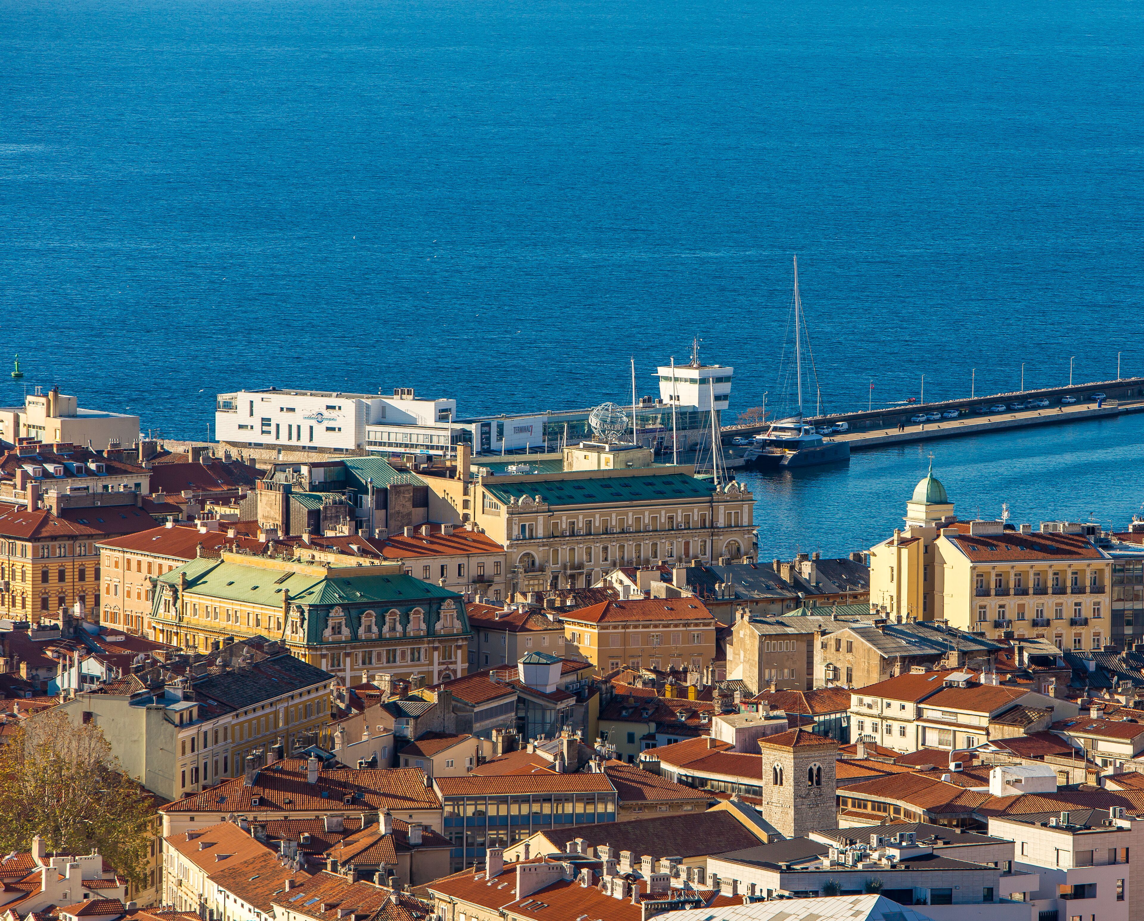 A coastal cityscape with historic buildings, a prominent tower, and a marina with boats.