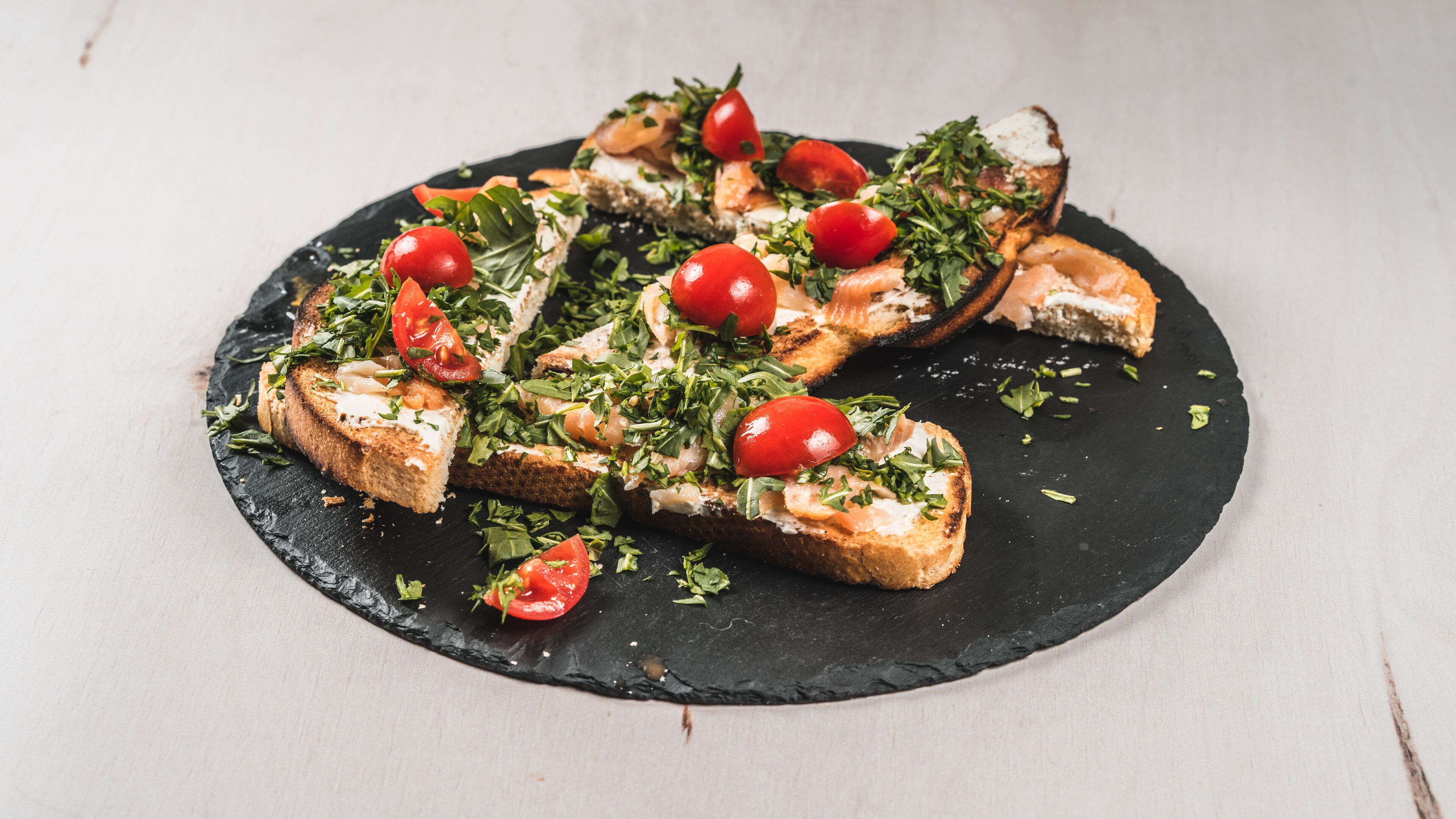 A black slate serving board with toasted bread topped with greens, tomatoes, and a creamy spread.