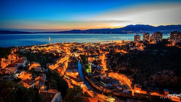 Un paisaje urbano costero al anochecer, con calles iluminadas, un puente destacado y montañas al fondo.