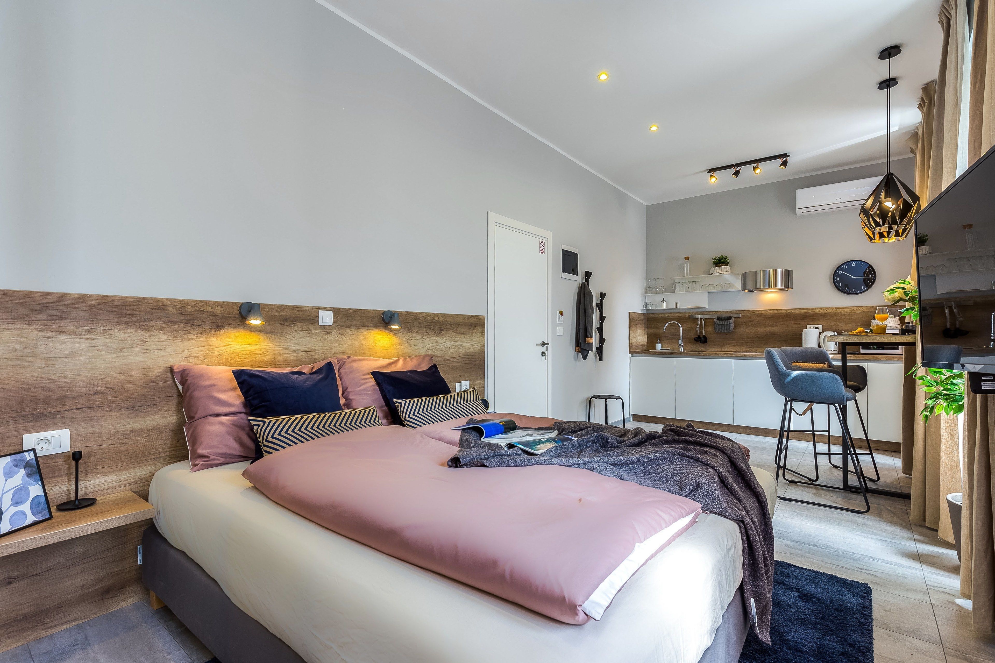 A modern bedroom with a large bed, bedside tables, a desk, and a kitchenette.