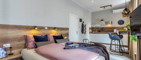 A modern bedroom with a large bed, bedside tables, a desk, and a kitchenette.