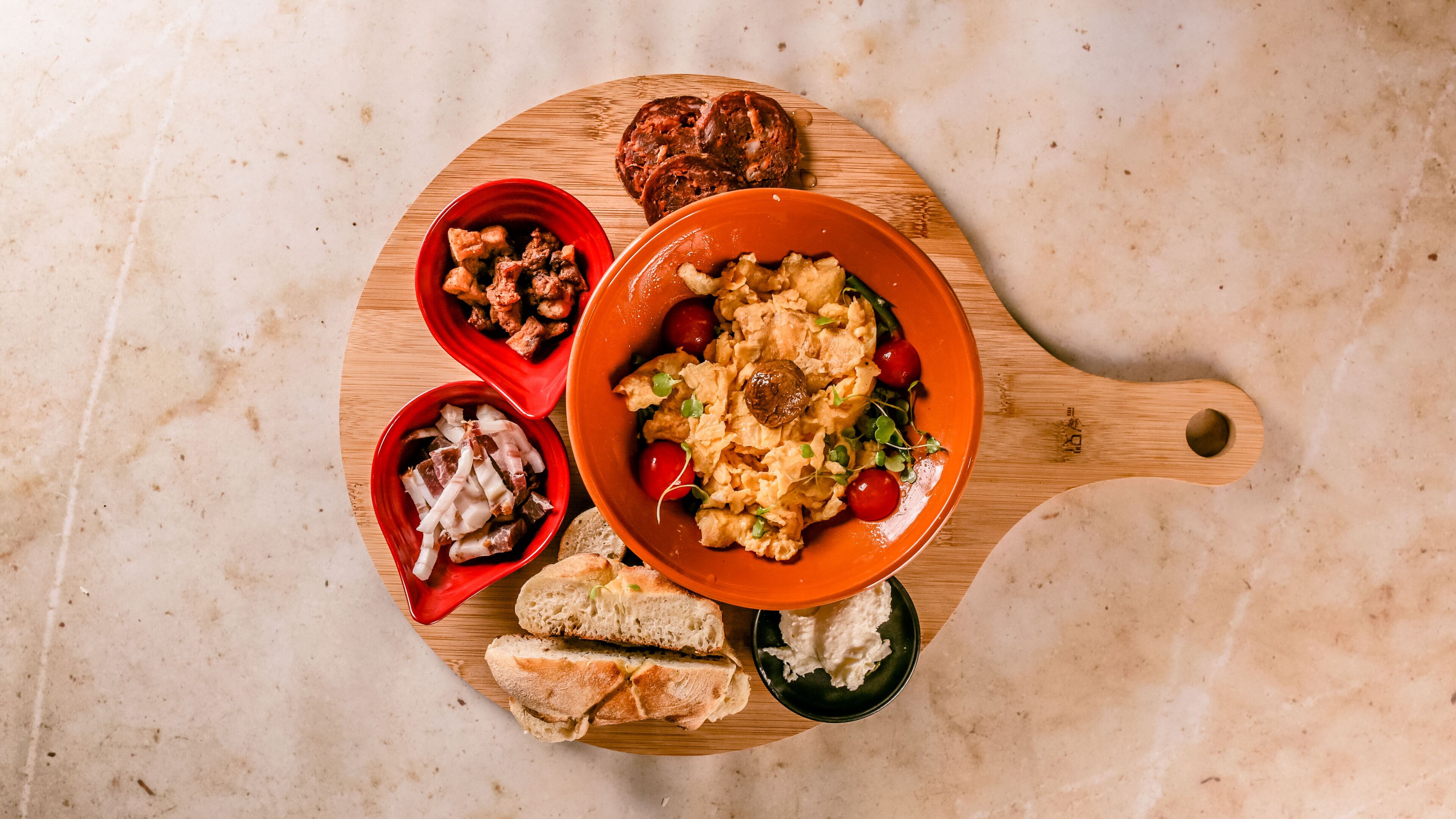 Una tabla de madera con diversos alimentos, incluyendo un tazón de huevos revueltos, un tazón de carne, un tazón de encurtidos y rebanadas de pan.