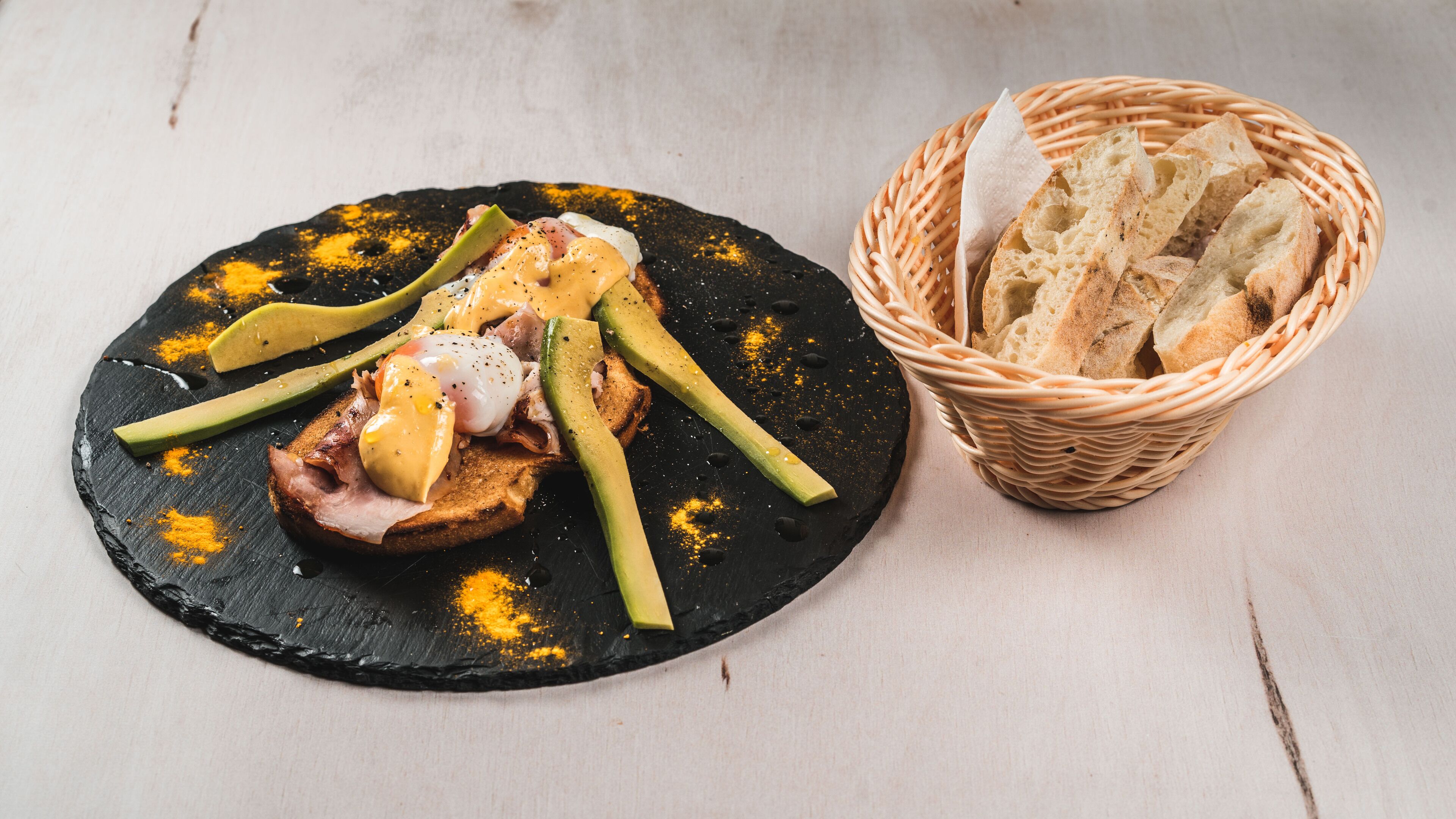A black slate serving board with a breakfast dish, including toast, eggs, and avocado, and a basket of bread.