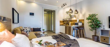 Une chambre d’hôtel moderne comprenant un lit, un canapé, une petite table avec une plante, un coin cuisine et une table à manger avec des tabourets.