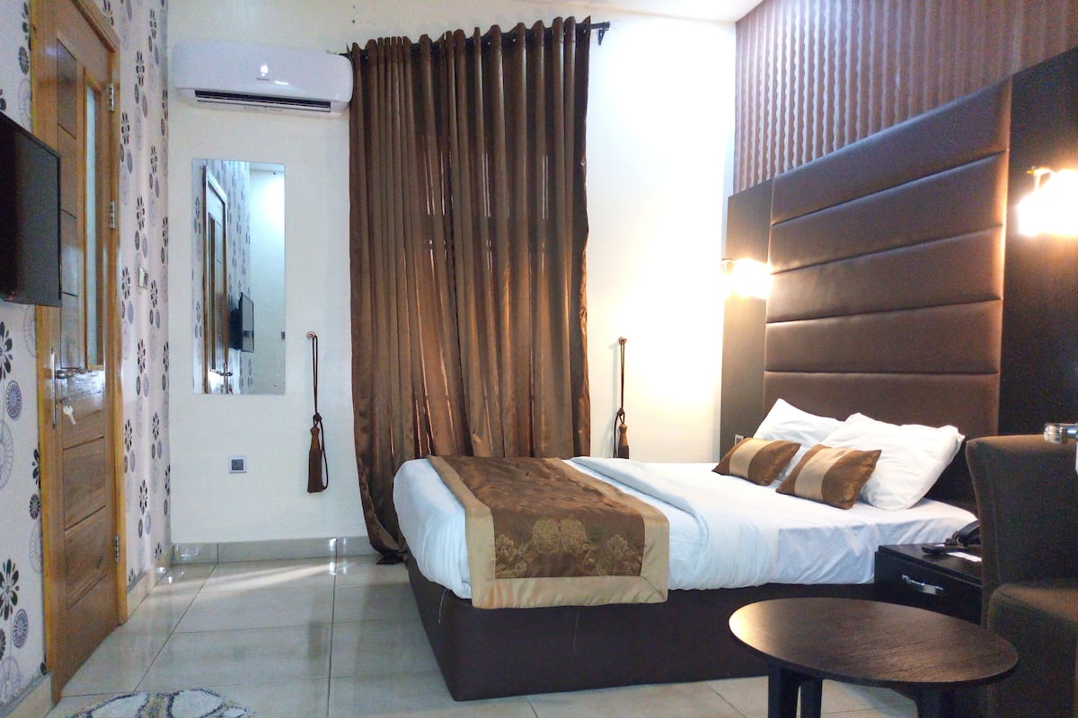 A hotel room with a bed, a chair, a table, a mirror, and an air conditioning unit.
