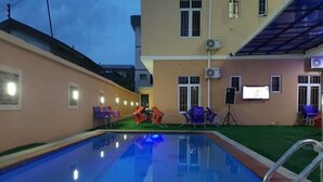 A swimming pool with a metal ladder, surrounded by a building with multiple windows and outdoor seating. - Bana Hotel & Suites (Lagos)