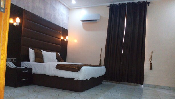 A hotel room with a large bed, a bedside table, an air conditioning unit, and a curtain. - Bana Hotel & Suites (Lagos)