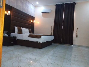 A hotel room with a large bed, a bedside table, an air conditioning unit, and a curtain. - Bana Hotel & Suites (Lagos)
