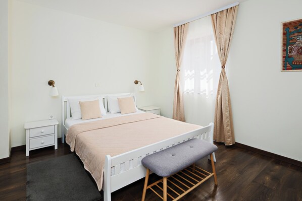 Un dormitorio con cama blanca, una banca, una mesita de noche, una lámpara y una ventana con cortinas.