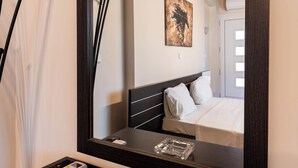 Hotellihuone, jossa on suuri peili, yöpöytä, jossa on kaukosäädin ja lasinen tuhkakuppi, valkoisilla lakanoilla varustettu sänky ja seinällä oleva taulu.