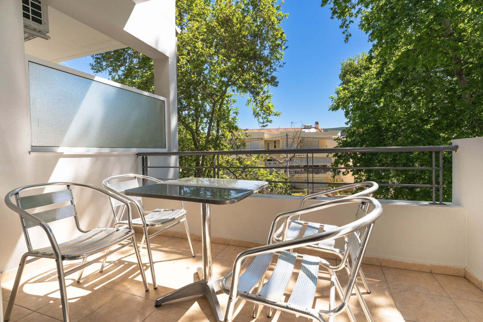 A balcony with metal chairs and a table, overlooking a residential area with trees and buildings.