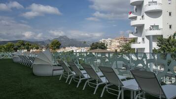 A rooftop terrace with rows of chairs and tables, a white building with balconies, and a mountainous backdrop.