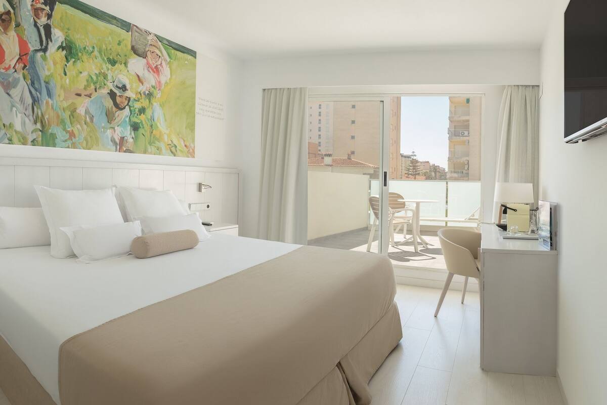 Habitación de hotel con una cama grande, un escritorio, una silla y un balcón con vistas a edificios.