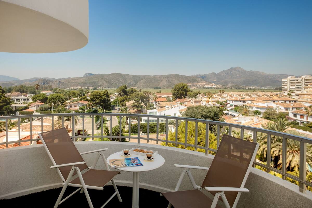 Un balcón con dos sillas y una mesa, con vistas a un pueblo y montañas a lo lejos.
