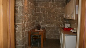 Cozinha compacta com parede de pedra, mesa de madeira com cadeiras, máquina de lavar e bancada com escorredor vermelho e chaleira.