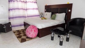 A bedroom with a bed, a desk, a chair, a pink ottoman, a lamp, and a suitcase. - De Fellas Palace Hotel & Suites (Ibadan)