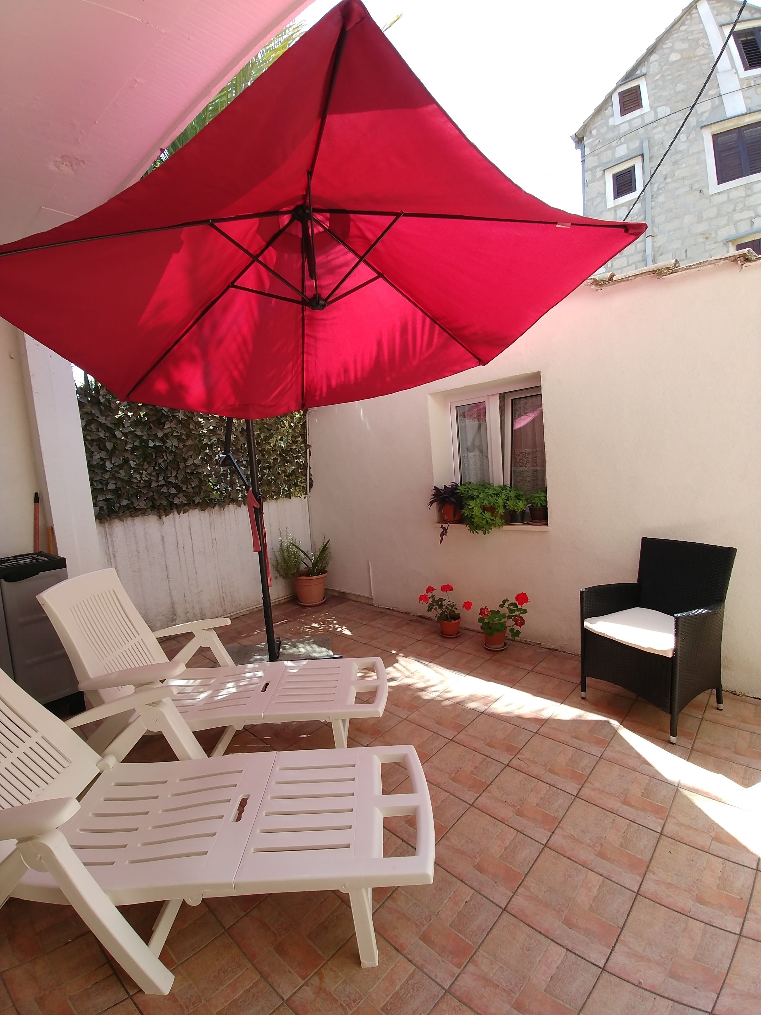 A patio area with two white plastic chairs, a red umbrella, and potted plants.