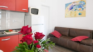 Eine moderne Küche mit roten Schränken, einer Mikrowelle und einem Schneidebrett mit Gemüse. Ein braunes Ledersofa mit roten Kissen und ein Bild an der Wand sind ebenfalls zu sehen.