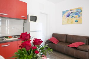 A modern kitchen with red cabinets, a microwave, and a cutting board with vegetables. There is a brown leather sofa with red cushions and a painting on the wall.