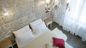 A bedroom with a stone wall, a bed with white bedding, a red pillow, a chandelier, and a nightstand. - Cool Place (Split)