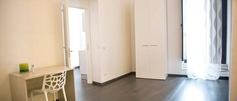 Una habitación con escritorio, silla, armario y ventana con cortinas translúcidas.
