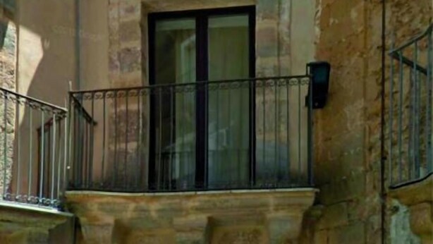 Un edificio de piedra con una puerta de entrada oscura, un balcón con barandillas metálicas y una puerta verde a la derecha.