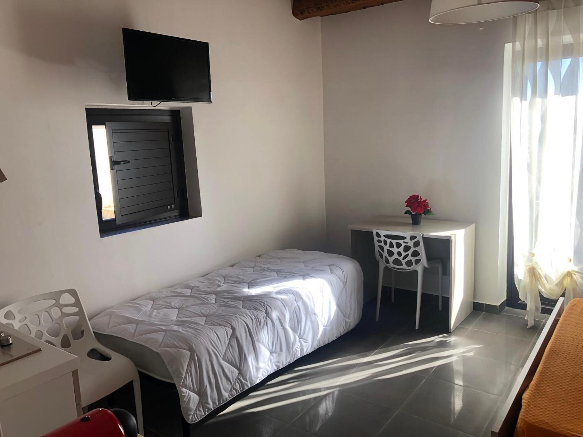 A small, well-lit room with a bed, a desk, a chair, and a TV mounted on the wall.