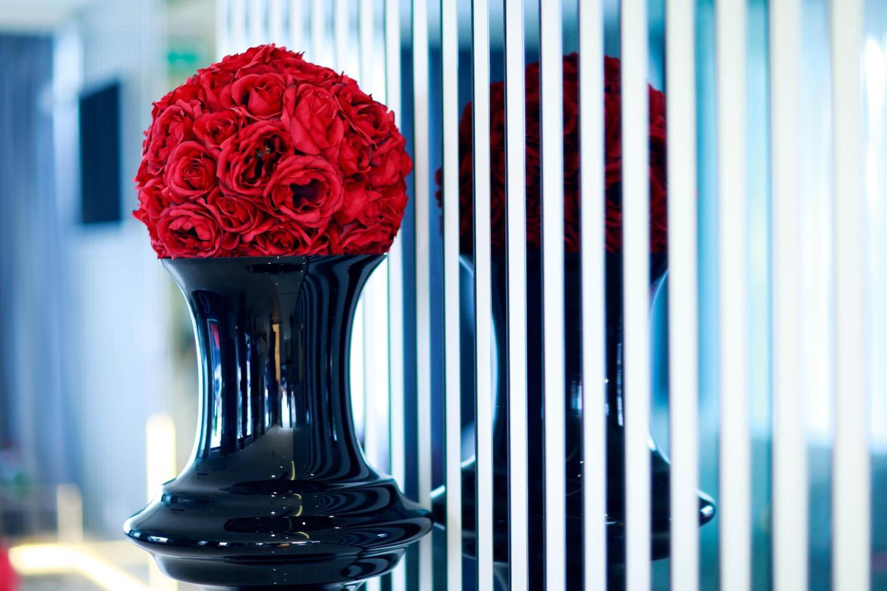 Un florero negro con un ramo de rosas rojas sobre una superficie reflectante, con líneas verticales en el fondo.