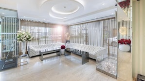 Une chambre d’hôtel moderne dotée d’une cloison en verre, de deux canapés blancs capitonnés, d’une table basse en verre et d’une grande fenêtre ornée de rideaux légers.