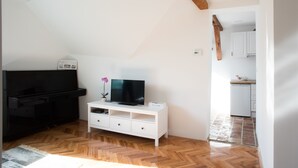A room with a wooden herringbone floor, a white TV stand with a flat-screen TV, a black piano, and a small white cabinet. - Sarajevo from A to Z (Sarajevo)