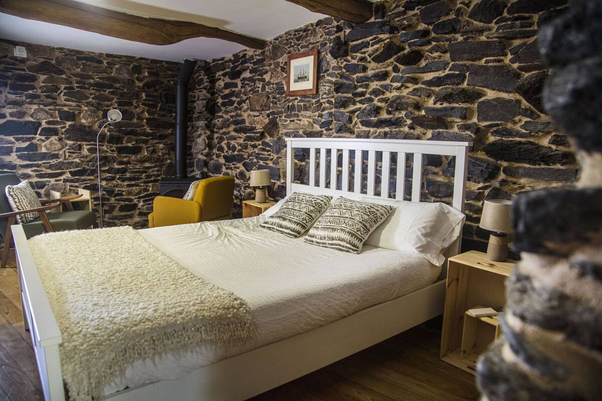 Ein Schlafzimmer mit Steinwand, einem weißen Bett mit gemusterten Kissen, einem gelben Sessel und einer Stehlampe.