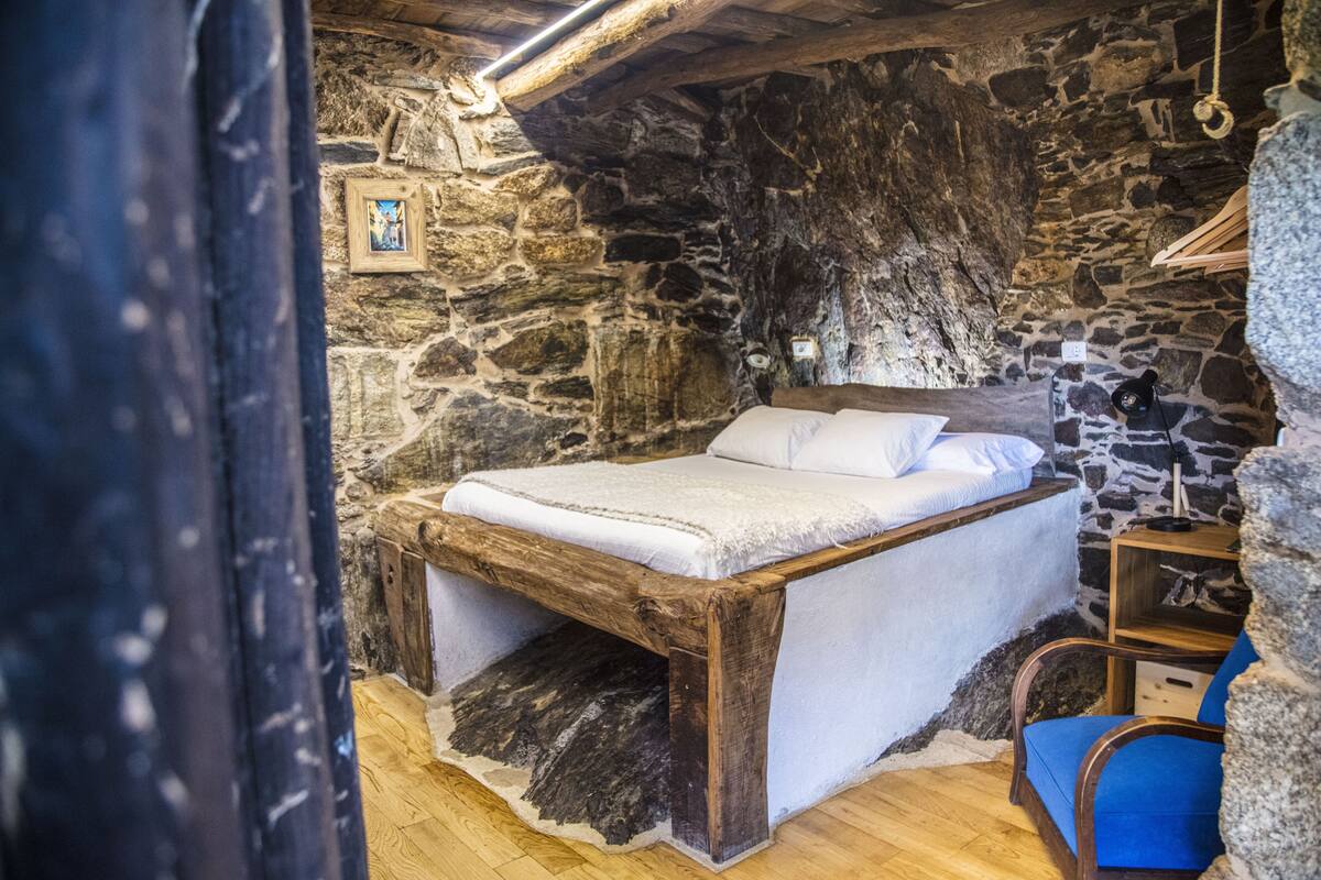 Ein rustikales Schlafzimmer mit Steinwänden, einem hölzernen Bettgestell, einem blauen Stuhl und einem kleinen Nachttisch.