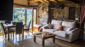 A cozy living room with a stone wall, wooden furniture, and a large window offering a view of the outdoors. - Cabo Do Mundo Casa Rural (Chantada)