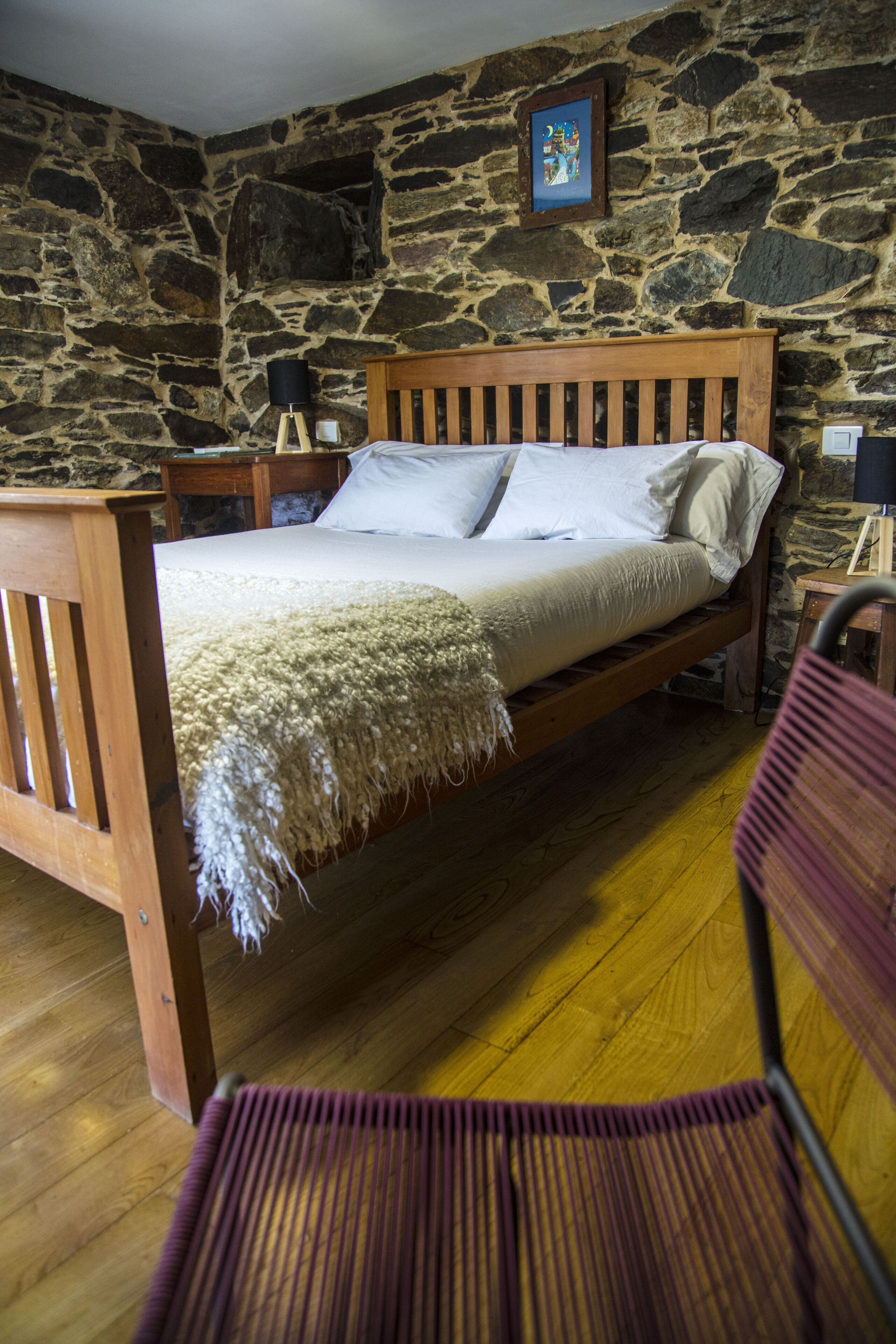 A bedroom with a wooden bed, stone walls, a wooden floor, and a chair.