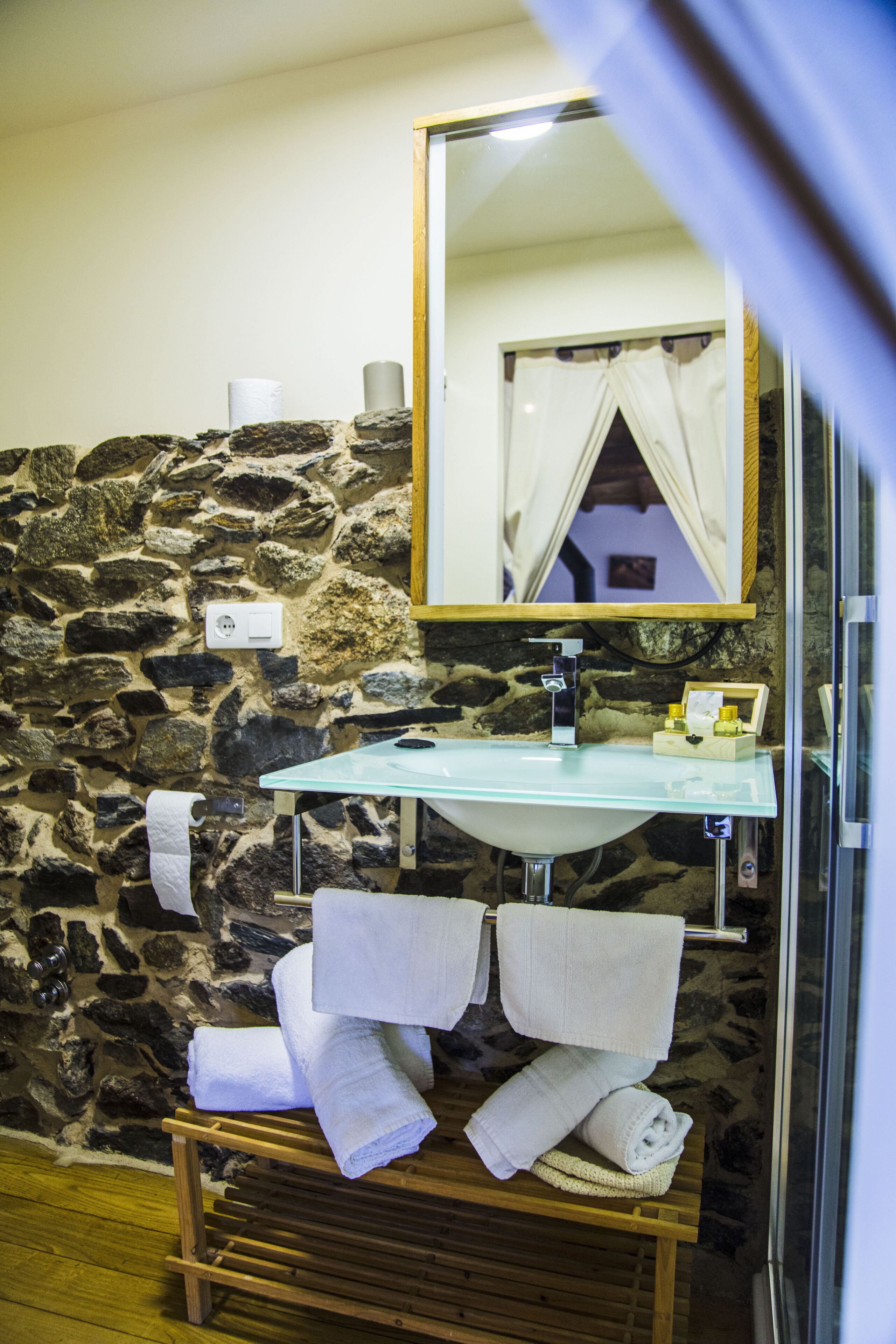 A bathroom with a stone wall, a glass sink, a mirror, and towels on a rack.