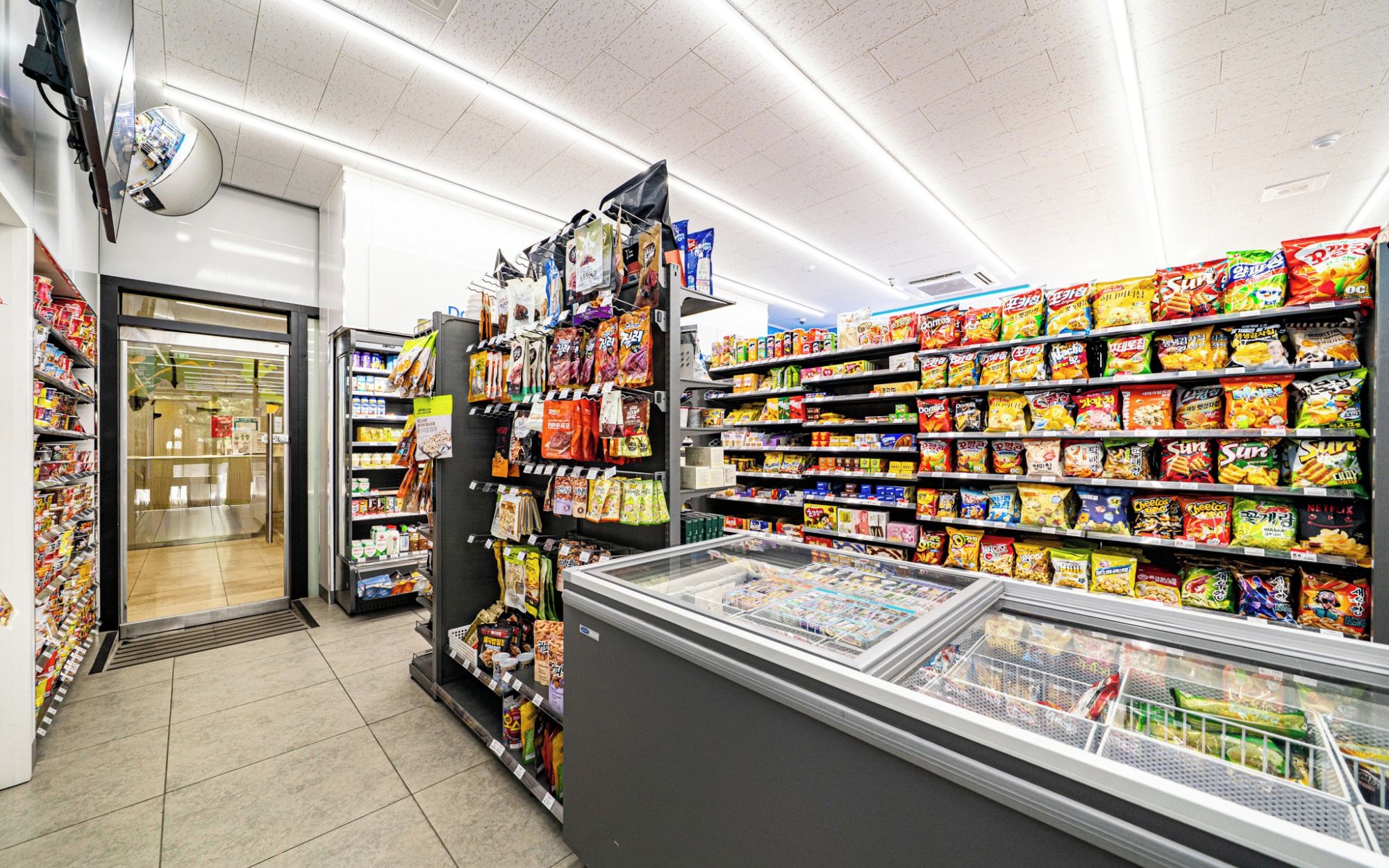 Un pasillo de un supermercado con diversos productos de comida rápida en estantes y una sección refrigerada al frente.