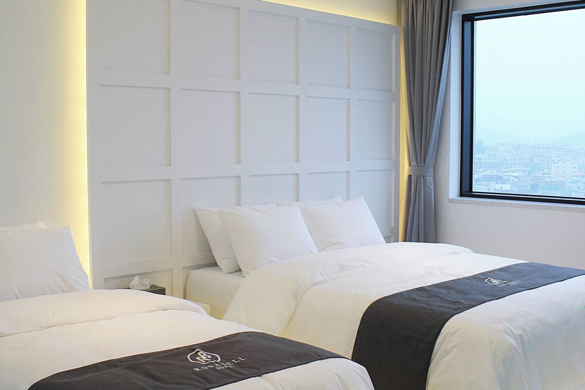 A hotel room with two beds, a large window with a city view, and a modern ceiling design.