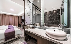 Moderni hotellihuone, jossa on suuri sänky, pesuallas ja peili, sekä suuri peili ja tummalla laatoituksella verhoiltu seinä.